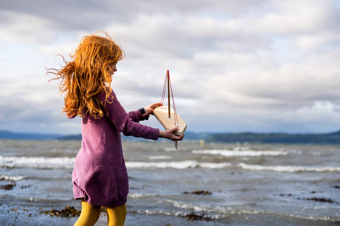Jente med lilla kjole holder en lekebåt opp mot havet i bakgrunnen
