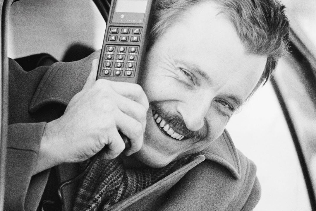 Mann stikker hodet ut av bilvindu i 1987. I hånden holder han en av datatidas store og klumpete mobiltelefoner.