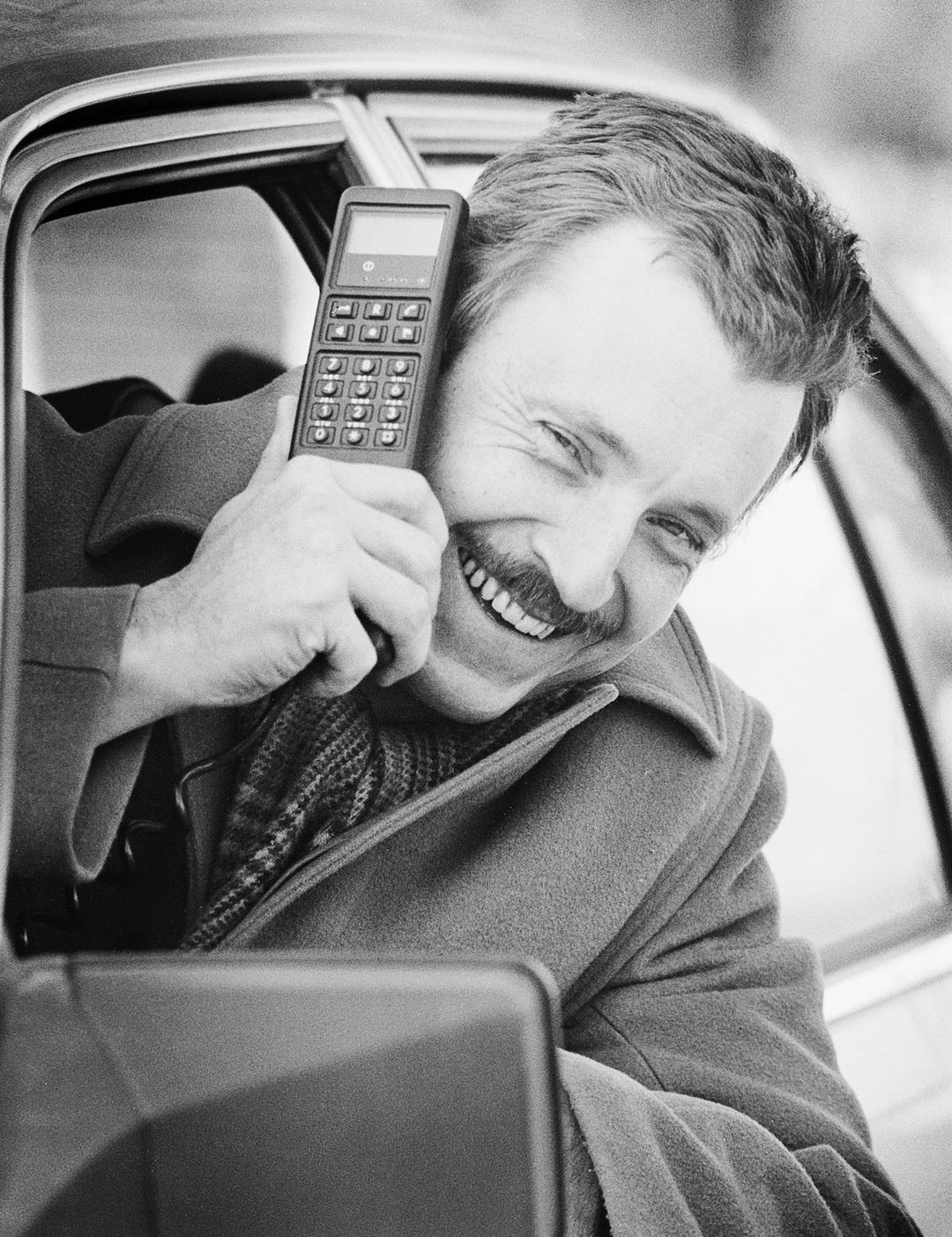 Mann stikker hodet ut av bilvindu i 1987. I hånden holder han en av datatidas store og klumpete mobiltelefoner.