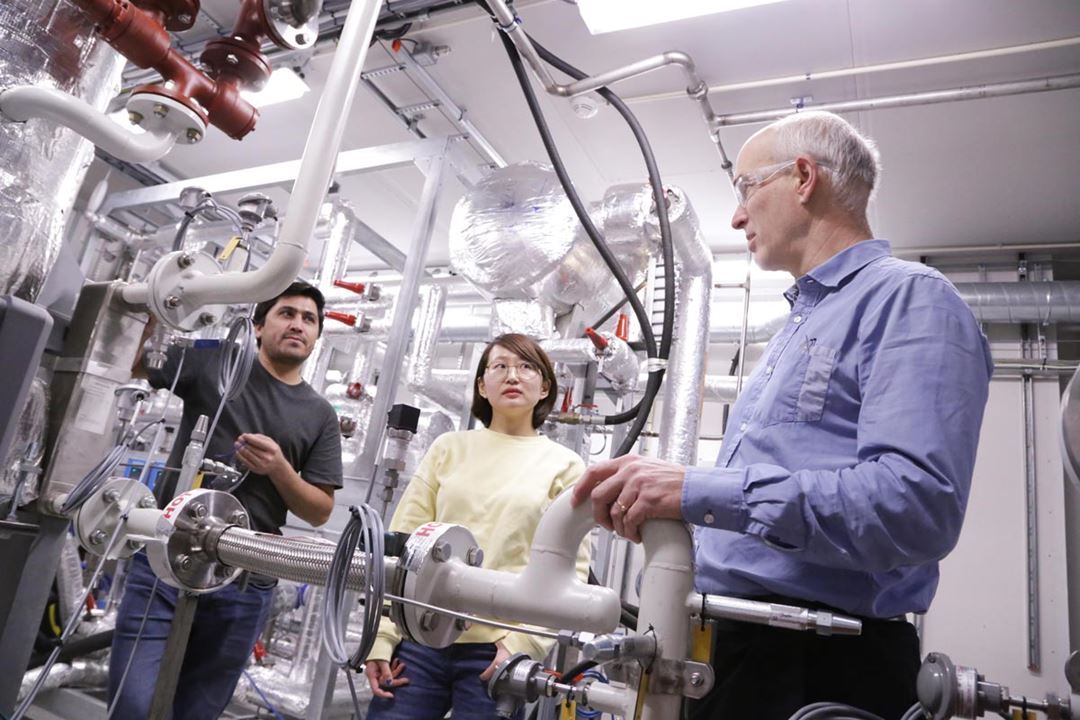 Tre personer står sammen i et teknisk laboratorium fylt med rør, ventiler og isolerte maskiner.