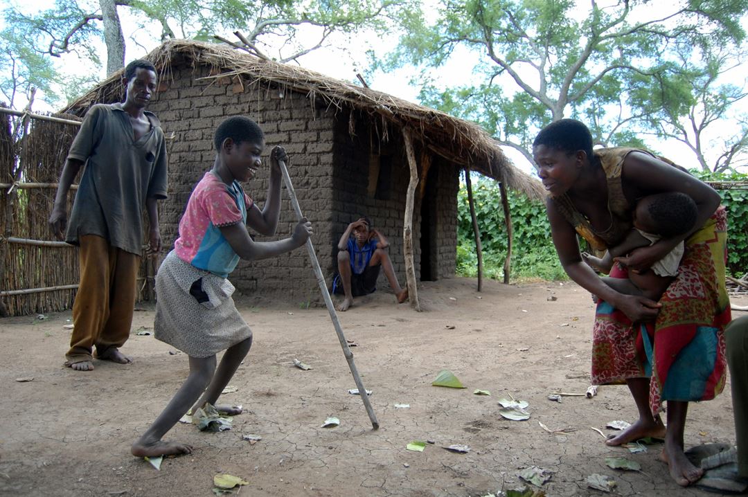  Funksjonshemmet jente i Afrika går over gårdstun, støttet til en lang stokk, mens familiemedlemmer oppmunter henne.