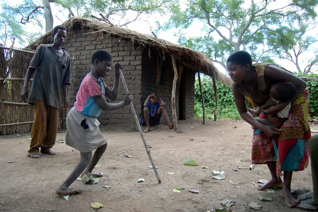 Funksjonshemmet jente i Afrika går over gårdstun, støttet til en lang stokk, mens familiemedlemmer oppmunter henne.