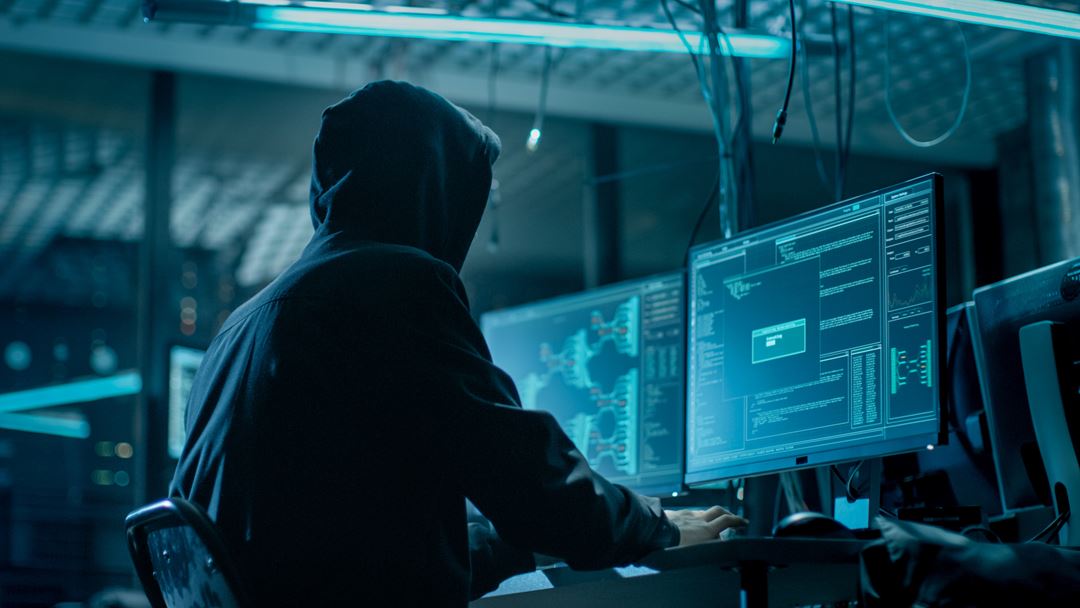 En mann med hette sitter foran en dataskjerm i et mørkt rom