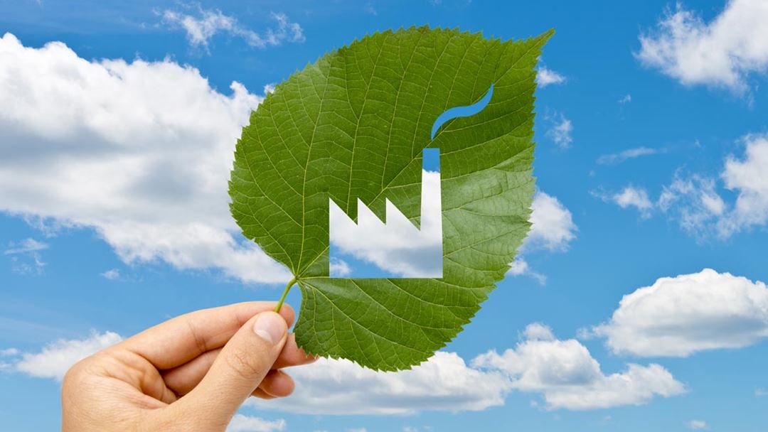Hand holding a leaf that has been cut out to represent a factory, against a sky background.