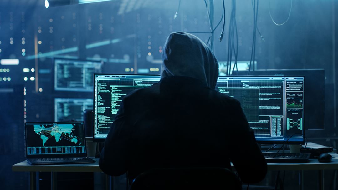 Mann med hette foran dataskjermer i mørkt rom