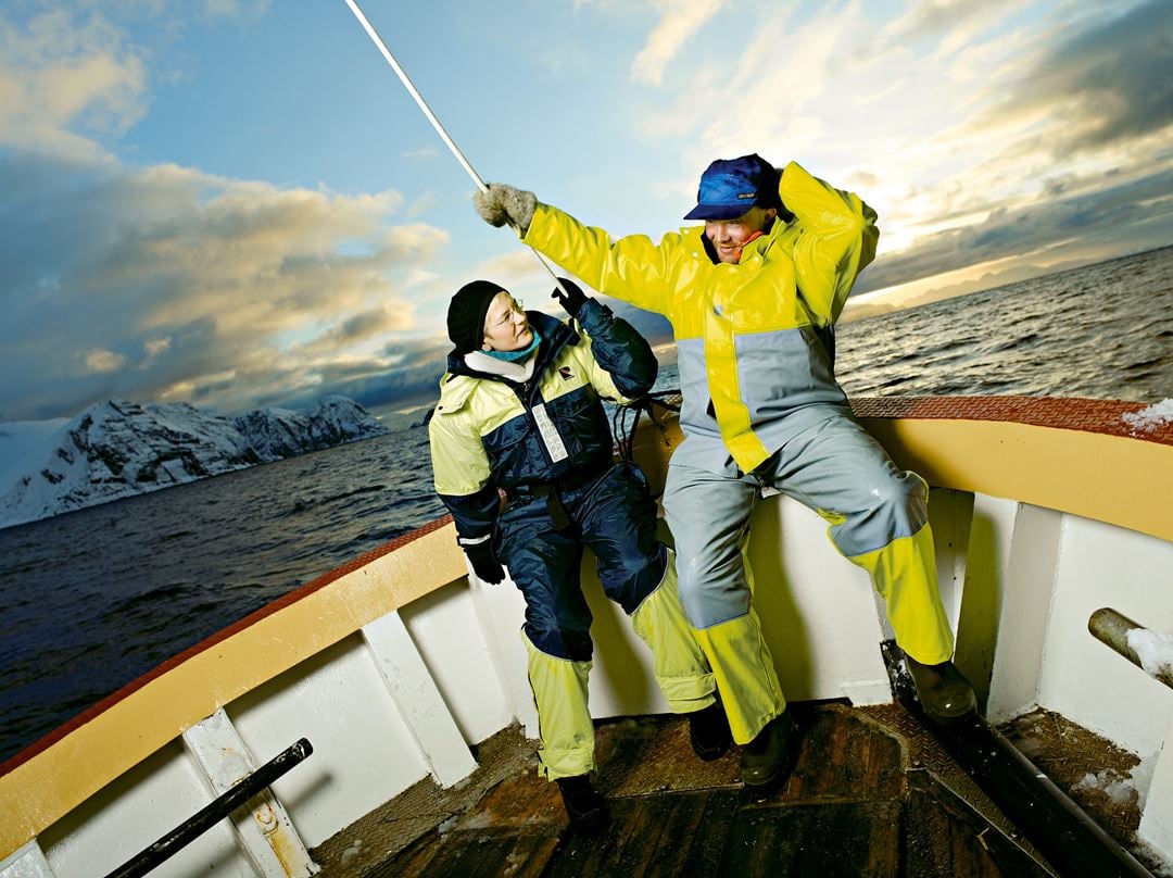 Mannlig yrkesfisker sitter foran i fiskeskøyte sammen med kvinnelig forsker. Begge kledt i gult og grått vanntett arbeidstøy.