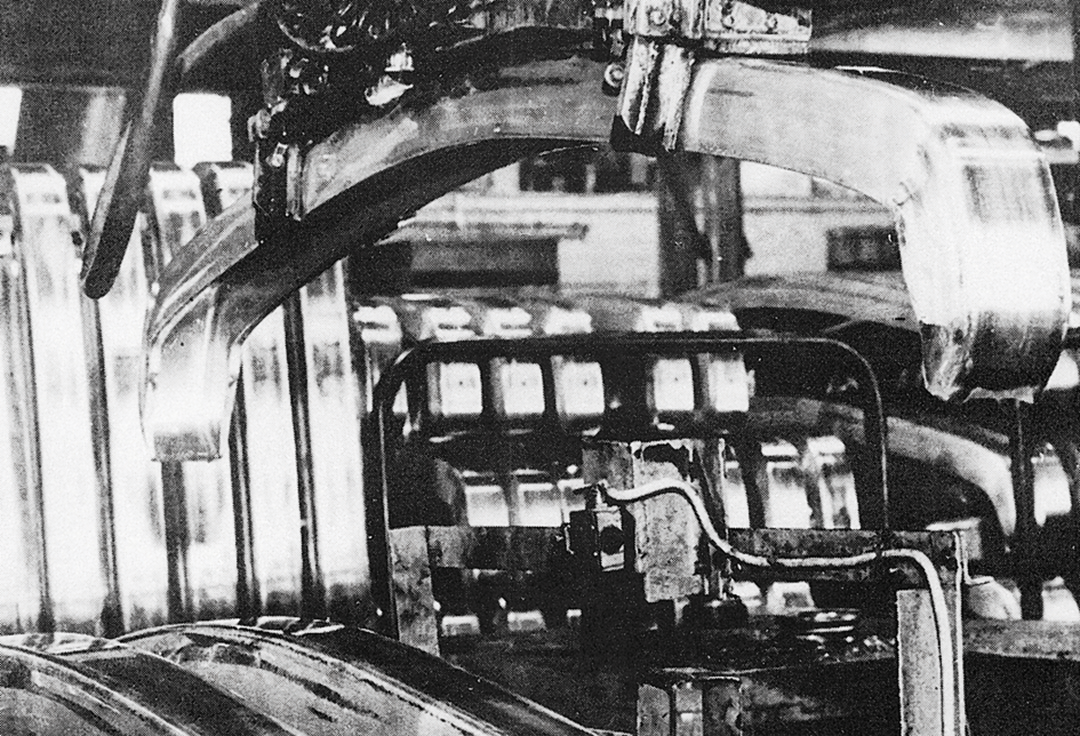 En kran heiser opp en nyprodusert støtfanger fra en rekke med tilsvarende produkter. Bildet er i svart hvitt, og er tatt i en hall hos Raufoss Ammuniksjonsfabrikker tidlig på 1970-tallet.