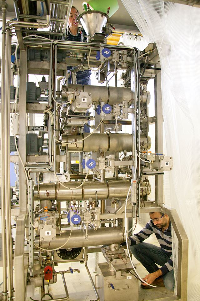 Med sin grønne bøtte laster senioringeniør Willy Georg Horrigmo inn oppkappede greiner og topper på toppen av laboratoriereaktoren, mens forsker Roger Khalil venter på resultatene i bunnen av prosessanlegget. En lang vei å gå for råstoffet. Til gjengjeld foredles det fra lavverdig til høyverdig biobrensel underveis. Foto: SINTEF / Gry Karin Stimo