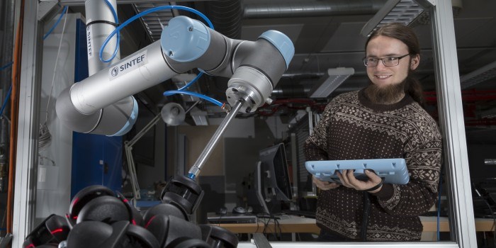 Forsker Sigurd Albrektsen har rigget seg til med pc'en rettet mot oppsettet av to robotarmer og en kasse med hjul. Her kan griperoboten plukke hjulene på fire ulike måter. Foto Thor Nielsen/SINTEF.