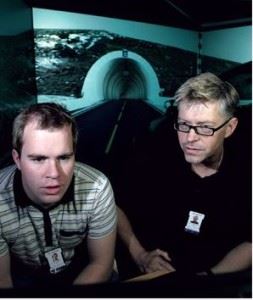 To menn menn sitter foran datautsyr i forkant av store videoskjermer som viser åpningen til veitunnel.