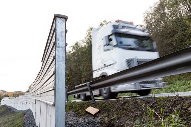 Lastebil passerer autovern, som litt bakenfor seg har en tradisjonell støyskjerm av tre.