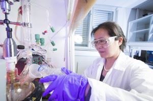 Dame i hvit lab-frakk, med vernebriller og fiollett arbeidshanske, arbeider med glasskkolber på kjemi-lab.