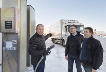 Tre menn står foran bensinliknende pumpe med hvit overbygd lastebil i bakgrunnen.