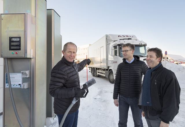 Tre menn står foran bensinliknende pumpe med hvit overbygd lastebil i bakgrunnen.