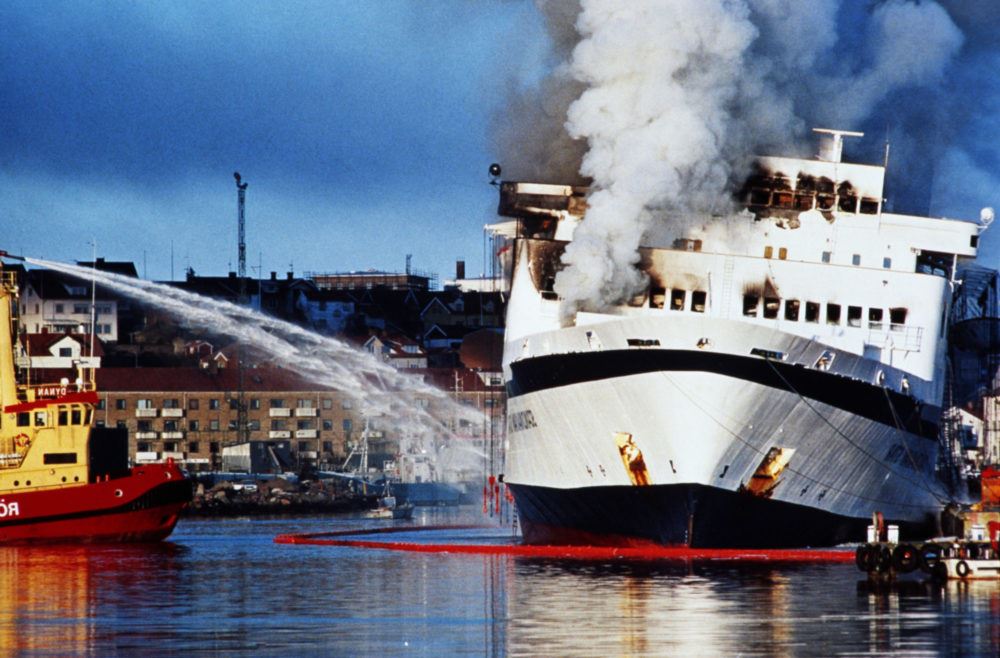 Røyk velter ut av Scandinavian Star idet den ligger ved kai i Sverige, og en sjøbrannsprøyte prøver å slokke brannen.