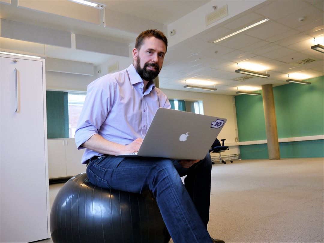 Mann siter i uformelt kontorlandskap med laptop.