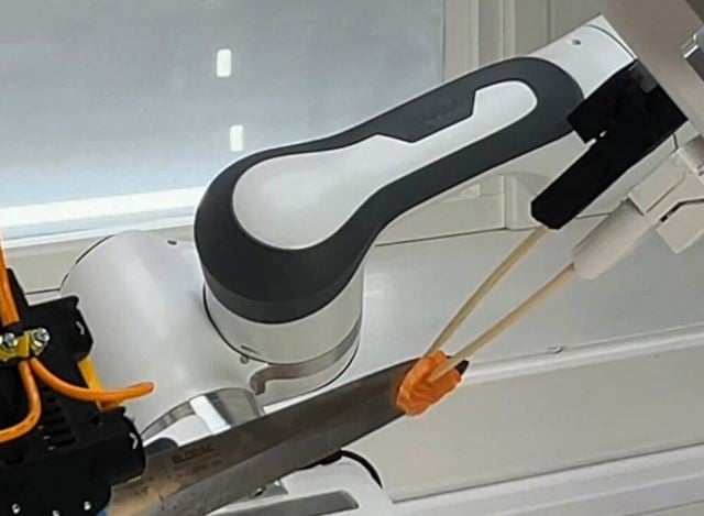 Robot with chopsticks removing a piece of salmon from a knife held by another robot arm